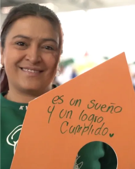 Mujer de 38 años sonríe feliz sosteniendo una casa de cartón de color naranja que contiene el texto "Es un sueño y un logro cumplido" gracias al proyecto de Viva mi casa líder en viviendas en Antioquia.